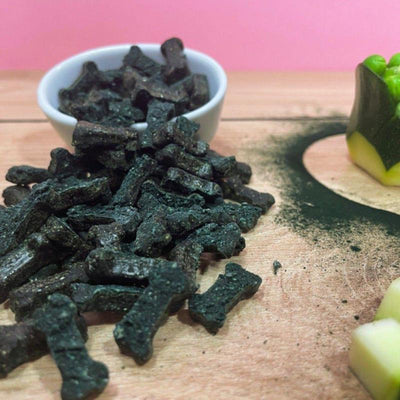 Petites friandises vertes en forme d'os à base de spiruline, accompagnées de courgettes, petits pois et spiruline en poudre sur une table en bois
